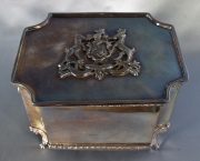 Cofre de metal plateado con escudo en la tapa. Alto: 10 cm. Frente: 17 cm.