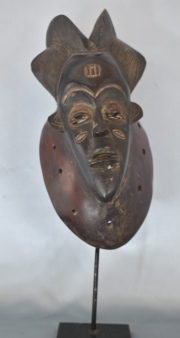 Máscara africana Guro, negra sobre fondo ocre. Escarificaciones y alto tocado. Alto 34 cm.