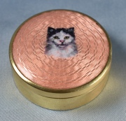 CAJA, de metal dorado con figura de gato de esmalte en la tapa, sobre fondo rosado.